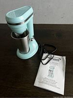 Light blue retro style milkshake maker with stainless steel cup and user manual on a dark surface. Power cord attached.