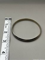 Silver bangle shown next to measuring tape, indicating diameter about 2.5 inches. Surface appears polished and smooth.