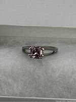 Close-up of silver tone ring with square-cut pink center stone placed on white cushion inside a box.