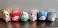 Six small Squishmallow plush toys lined up on a dark surface against a grey background. From left to right: rainbow unicorn, sushi roll, white and blue narwhal, lavender alpaca/llama, blue penguin, purple unicorn.