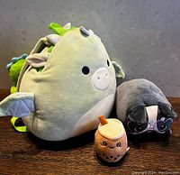 Shows three assorted Squishmallow plush items on a wooden surface: a green dragon backpack, a gray cat plush, and a bubble tea plush keychain.