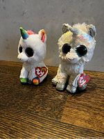 Two TY unicorn Beanie Babies placed on a wooden surface against a stone wall. One plush unicorn has rainbow-colored horn and details, the other is covered in iridescent sequins with silver horn and feet.