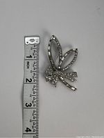 Silver tone bow brooch beside measuring tape showing about 2 inch size. Clear small stones and baguette stones on the bow shape. Some baguette stones missing.