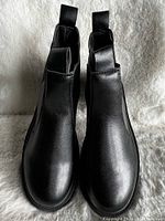 Front view of the pair of new women's black Chelsea boots on a white fur background, showing smooth toe and leather-like material.