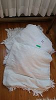 Three white chenille bedspreads stacked on a wooden floor, showing the fringed edges and basketweave texture.