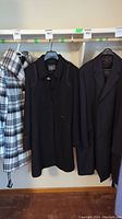 Three trench coat style coats hanging on rack showing black Berkley Worsted tailored coat in center and black Projek Raw buttoned coat on right side
