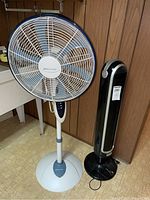 Image showing both a white and grey 40 inch Dynamo pedestal fan and a black 48 inch Bionaire tower oscillating fan side by side against a wood paneled wall.