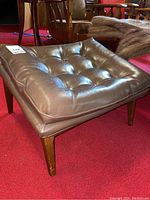 Front and side view of the brown vinyl tufted cushion on the wood framed ottoman showing slight surface wrinkles and wooden legs.