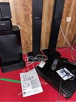Full view of the system components including tower speakers, subwoofer, remote, instruction manual, and cables on carpet