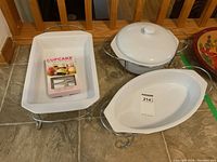 Three white stoneware dishes including rectangular, oval and casserole with lid, all with metal holders, displayed on a tiled floor and in front of wooden railings.