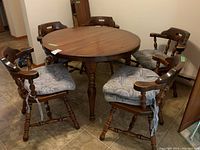 Round dining table surrounded by five wooden chairs with cushions, showing overall wear and scuffs.