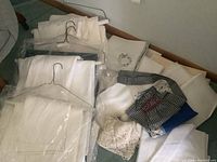 View of multiple sets of folded white linen tablecloths and napkins wrapped in plastic, laid on floor with some aprons on top, showing mix of fabric types and patterns.