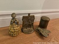 Photo showing four vintage brass items on the floor: a lady figurine bell, a house-shaped piece, a small cylindrical container, and a frog figurine.