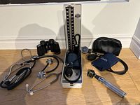 Full lot laid on a wooden floor showing all medical equipment including sphygmomanometer, blood pressure cuff, stethoscopes, otoscope, binoculars, and black pouch.