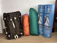 Photo showing a black backpack, two tents in carry bags (green and orange), and two large blue Airolife-branded foam sleeping mats upright against a door.