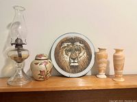 Photo showing 2 alabaster vases with cream and beige swirl pattern, 2 antique clear glass oil lamps, small brass and enamel urn with flower design, and a circular enamel lion medallion.