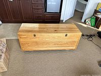 Closed wooden chest showing light wood grain and two metal clasps on the front, placed on carpeted floor in front of kitchen cabinetry.
