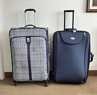 Two upright soft-sided suitcases side by side on carpeted floor. Left suitcase plaid fabric with four multi-directional wheels. Right suitcase navy fabric with two fixed back wheels, combination lock on top, and handle extended.