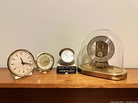 Five vintage clocks displayed on wooden surface, including Kieninger & Obergfell brass carriage clock, two travel clocks, and two bedside alarm clocks.