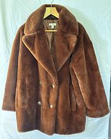 Brown teddy bear faux fur double-breasted jacket with lapels and pockets hung on wooden hanger against white background.