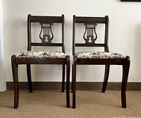 Photo of two straight backed wooden chairs showing carved lyre-shaped backrests and floral embroidered seats