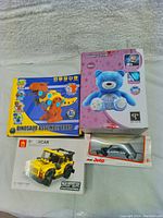 Four brand new toys shown in packaging: dinosaur assembly set, blue musical bear, yellow building blocks car, and a jeep model car
