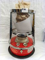 Front view of the portable kerosene heater stove showing red base, control knobs, and stainless steel mesh guard with paper inside.