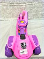 Front view of a pink kids scooter with purple handles and wheels, showing the smiley face sticker and manufacturer's logo.