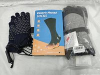 Photo of navy blue heated gloves, electric heated socks in box packaging, and black insulated heated gloves in plastic bag.