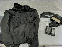 Black Venustas men's heated jacket laid out flat with accessories and packaging visible on a white surface.