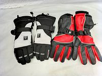 Two pairs of winter sports gloves, black/grey and black/red, showing front sides with adjustable wrist straps and materials details.