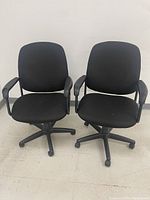 Photo showing two black office chairs side by side on a floor against a white wall. Chairs have fabric seats and backs, armrests, and wheeled bases.