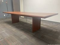 Solid wood rectangular boardroom table with two large panel legs shown from a side angle on carpeted office floor.