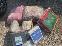 Lot items arranged outdoors on gravel with vehicle tire in background; visible are pink quilted blanket, floral blanket, Christmas blanket, grey towels, bedding, and mattress protector.