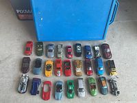 Top-down view of 24 assorted toy cars placed in rows on a gray surface with a blue carrying case behind them.