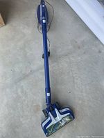 Full view of the blue Dirt Devil upright vacuum cleaner on concrete floor, showing the cleaning head and handle with power cord wrapped.