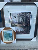 Two framed art prints leaning against a wall on a concrete surface. The larger piece shows a lake and forest scene with mountains.
