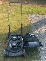 Full view of black Yard Machines 21-inch gas mower with attached handle and black fabric collection bag positioned on grass and gravel surface