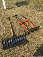 Photo of a large black plastic snow shovel with wooden handle placed on grass beside an orange manual lawn dethatcher with black wheels.
