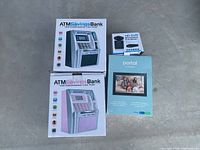 Four boxed items on concrete surface: two ATM Savings Banks (gray and pink), an HD DVR security camera, and a Facebook Portal video calling device.