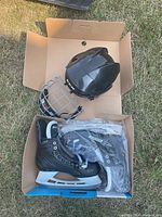 Top view photo showing the black hockey helmet with cage and Bauer ice skates packed in cardboard boxes on grass