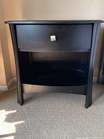 Front view of the black wood bedside table showing the drawer and lower open shelf.
