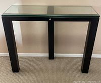 Front view showing the full console table with its black metal legs and clear glass top against a beige wall and carpeted floor.
