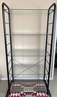 Front view of glass shelving unit with five clear glass shelves and black metal frame against a beige wall.