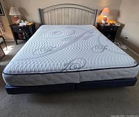 Full view of the king size mattress and black box spring on a carpeted floor with a steel bed frame headboard background. Two nightstands with lamps on each side.