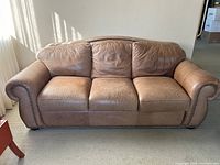 Front view of brown leather sofa showing three seats and rolled arms with nailhead trim, all cushions present with visible creasing and soft wear.