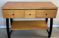 Front view showing three drawers with black knobs, wooden body with curved black metal legs, and lower wooden shelf.