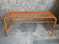 Orange wire bench against a textured wall, front and side view of entire bench, metal tubular legs and grid wire top surface.