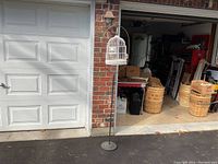 Full view of the antique wire domed bird cage on tall brass and iron stand placed outdoors in front of a garage and brick wall.