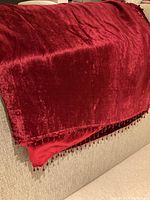 Burgundy throw fabric shown with velvety texture and beaded fringe along edges.